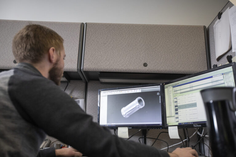 Person sits at computer, looking at a 3D model of a metal cylinder on the monitor.